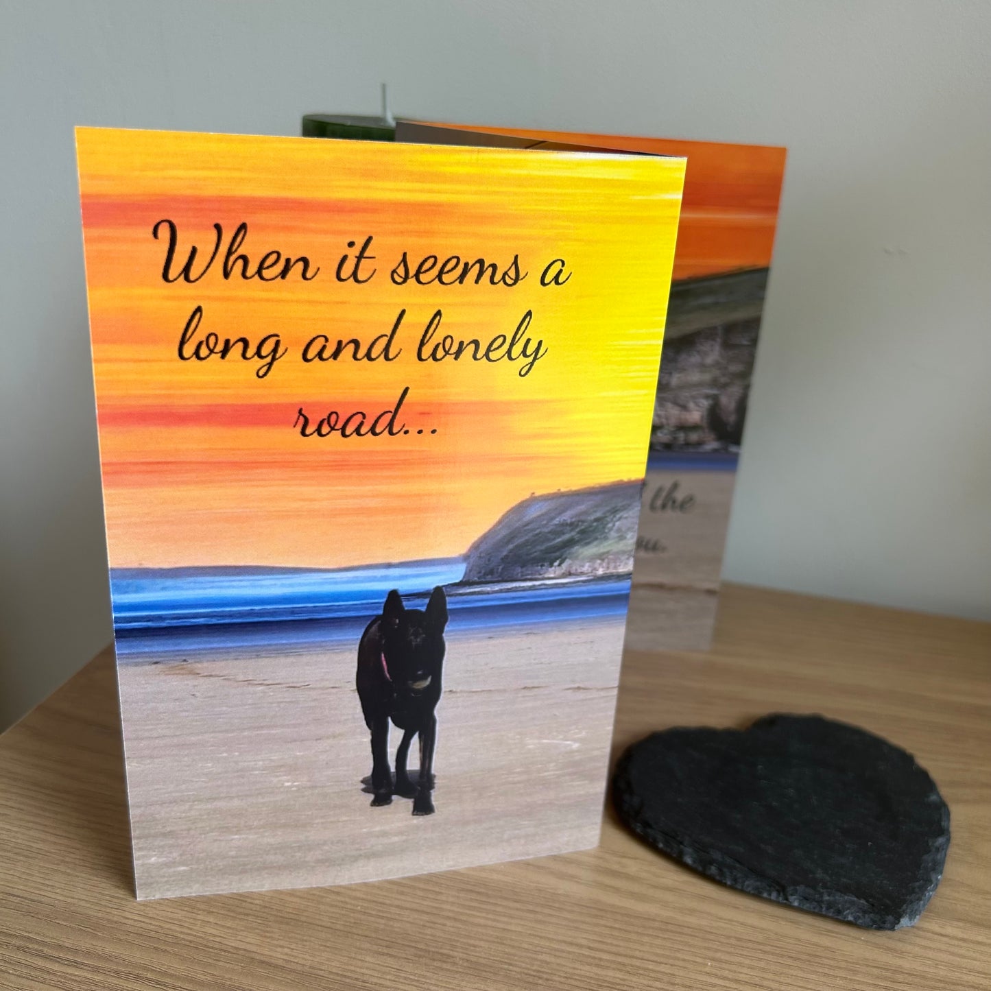The image shows a photo of the front of the Bare Cards Dog Photo Sentiment Card 'Lonely Road' showing a dog trotting up the empty beach with a sunset background and the words 'When it seems a long and lonely road'.