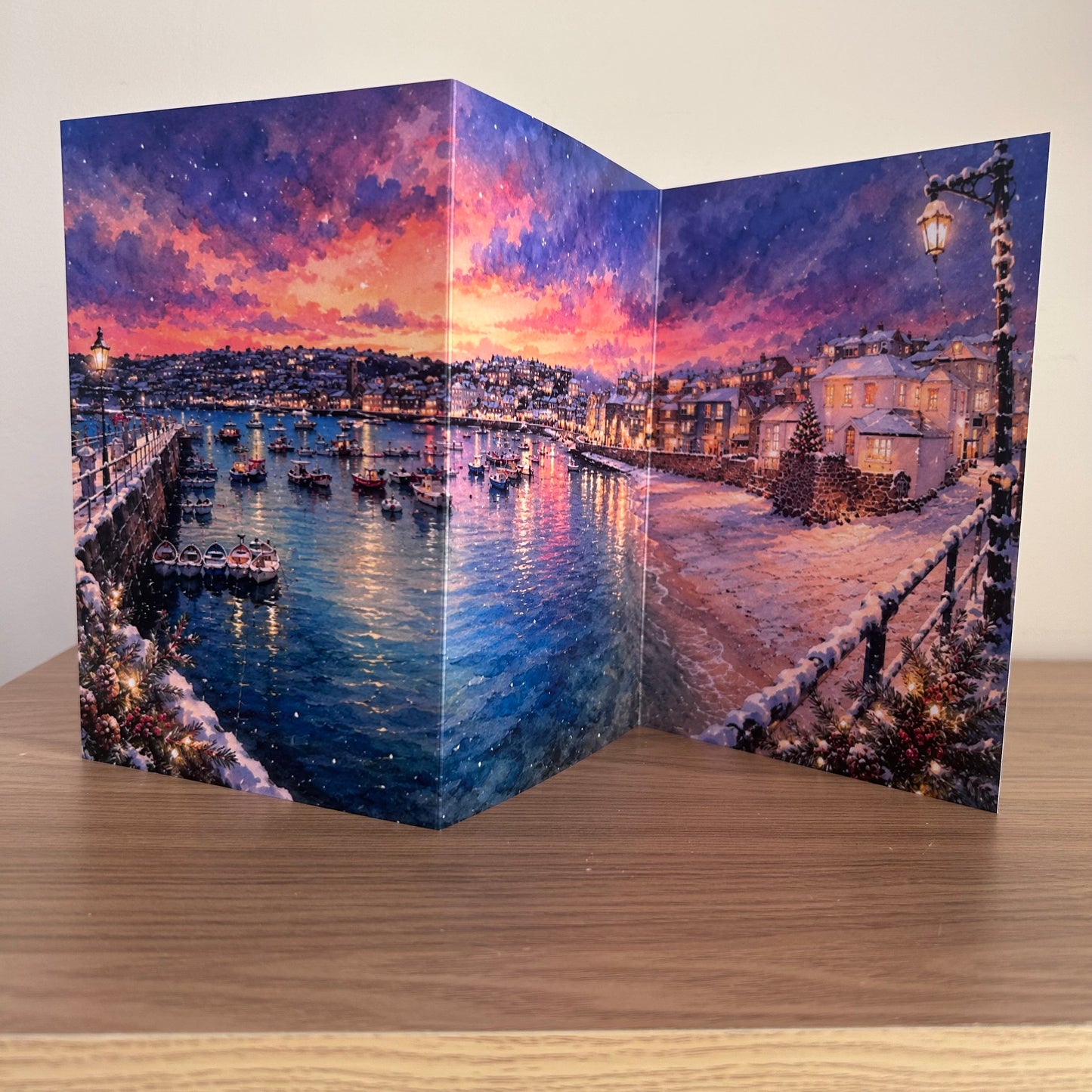 A trifold accordion-style greeting card standing open on a wooden table, printed with a colorful winter harbor scene at St Ives featuring boats, snowy streets, and a dramatic sunset sky.