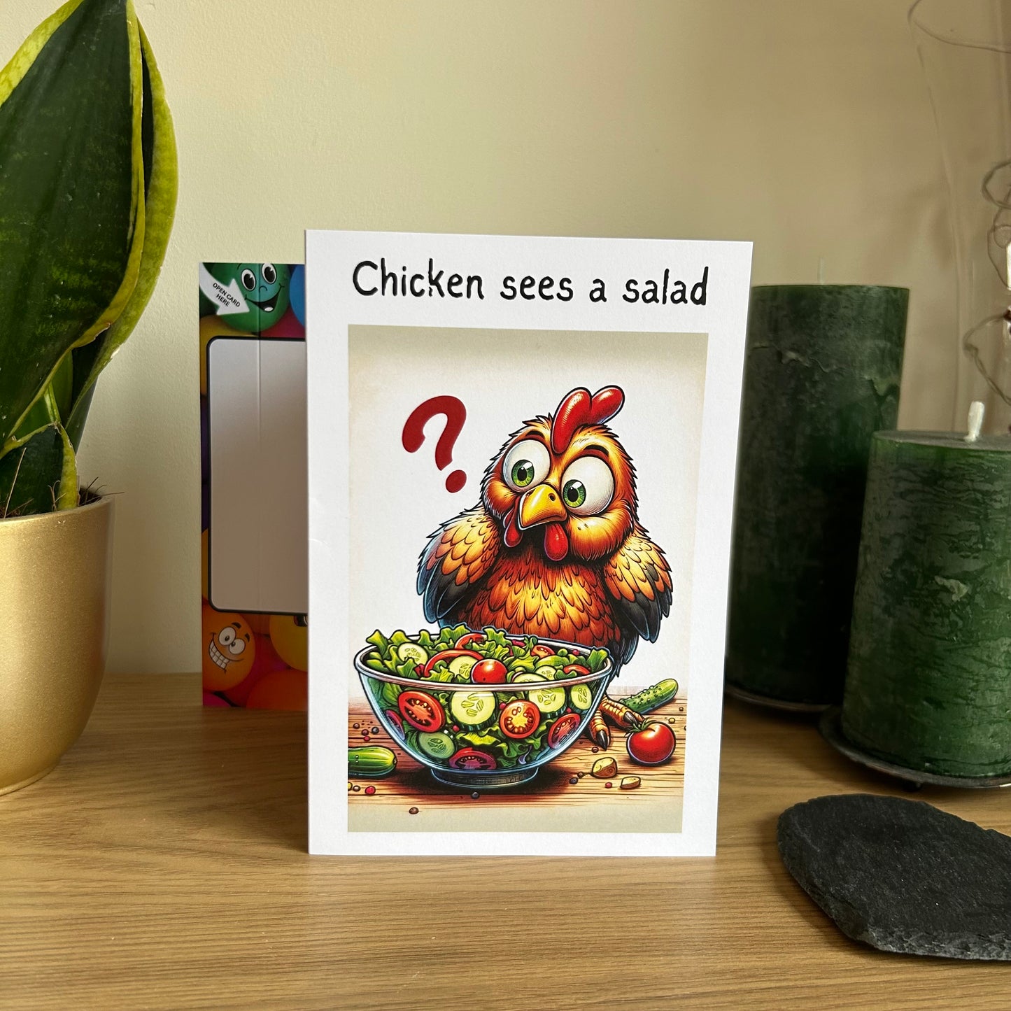 Image shows the Bare Cards Giggle Balls Humour Range 'Chicken Sees a Salad' Birthday Card displayed in a home setting. The illustration shows a startled cartoon chicken looking at a bowl of salad with a question mark hanging in the air and the caption 'Chicken sees a salad'.