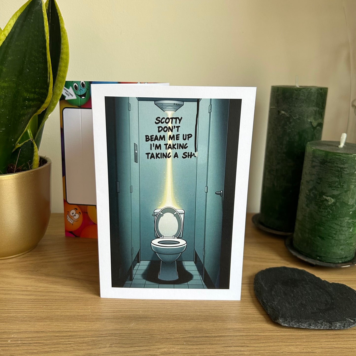 Image shows the Bare Cards Giggle Balls Humour Range 'Scotty Joke' Birthday Card displayed in a home setting. The illustration shows an empty public toilet cubicle with the toilet seat up and the words "Scotty Don't Beam Me Up, I'm taking a SH...." written on the wall with a marker pen sliding upwards suggesting that Scotty did beam them up mid toileting!