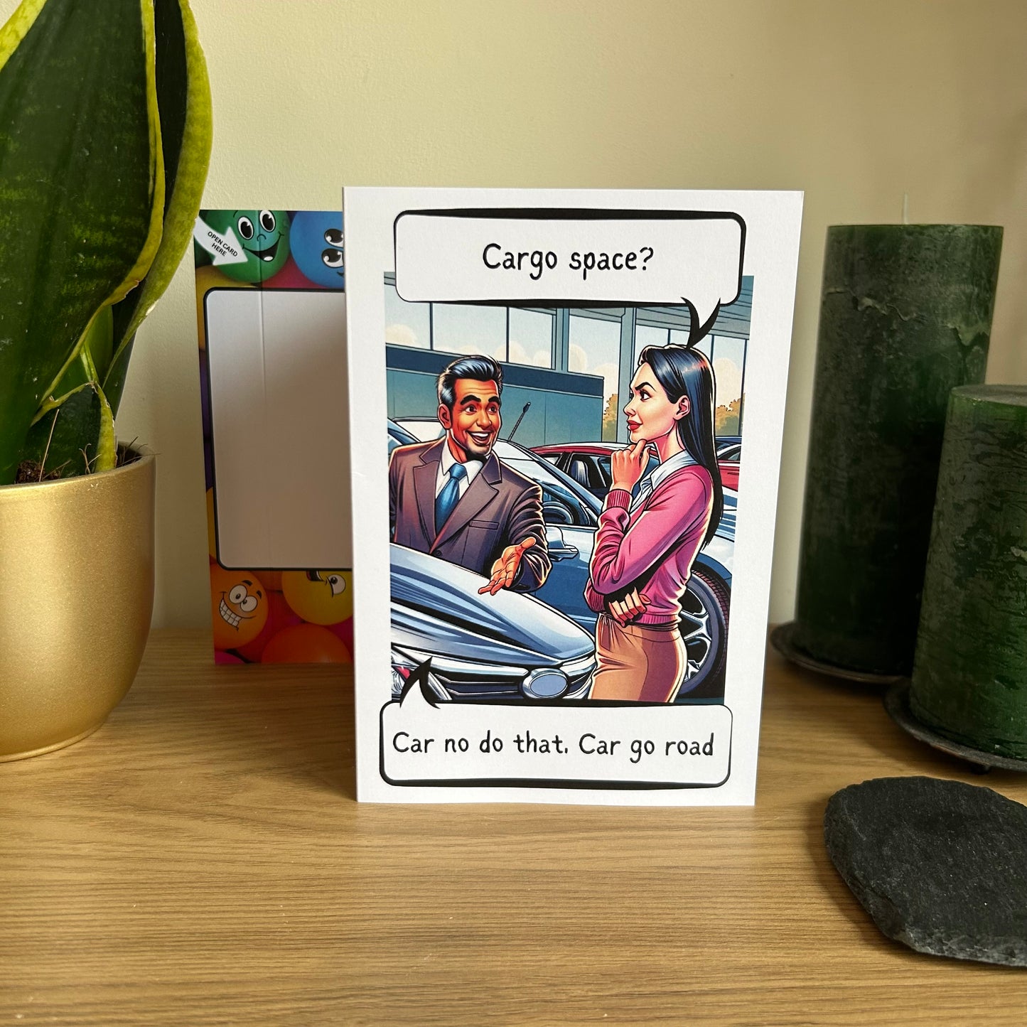 Image shows the Bare Cards Giggle Balls Humour Range 'Cargo Space' Birthday Card displayed in a home setting. The illustration shows a car salesman showing a pretty lady a car. She is asking the question 'Cargo Space?' and the salesman replies 'Car no do that, Car go road'
