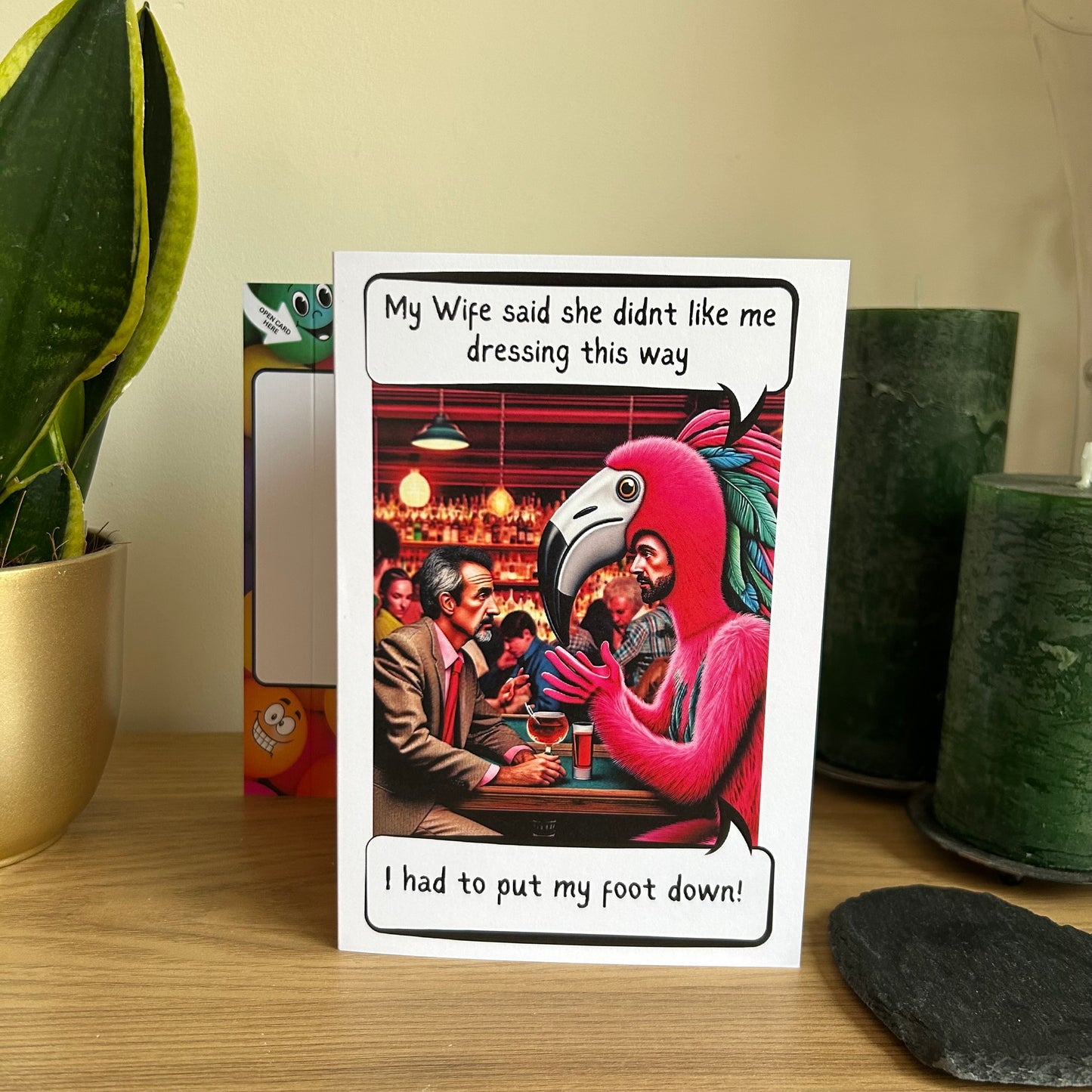 Image shows the Bare Cards Giggle Balls Humour Range 'Famingo' displayed in a home setting. The illustration shows two men sitting in a pub having a drink. One is dressed in a suit and the other in a bright pink flamingo costume. The caption reads 'My wife said she didn't like me dressing this way. I had to put my foot down!'