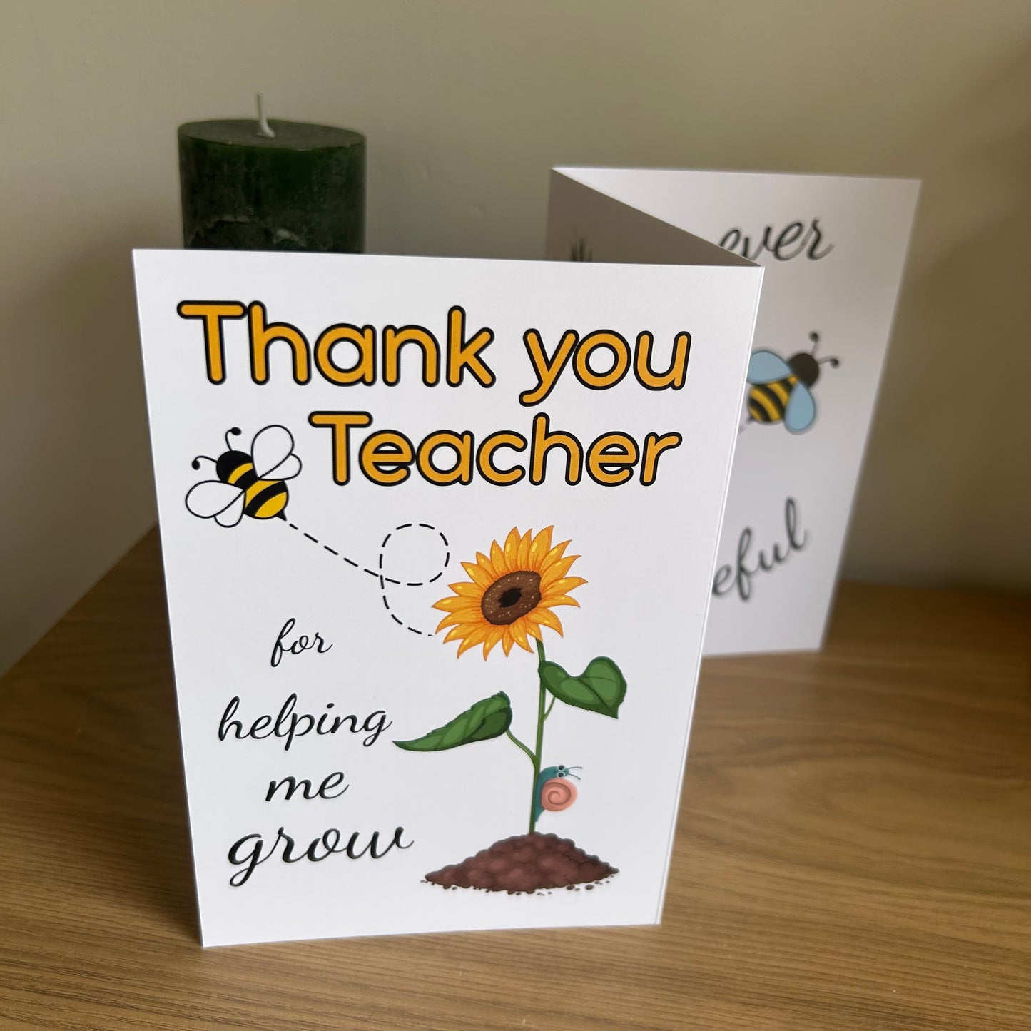 The photo shows the front of the Bare Cards Thank you teacher card with images of a sunflower and a bee and the caption "Thank you teacher for helping me grow". It is displayed on a shelf with a green candle behind.