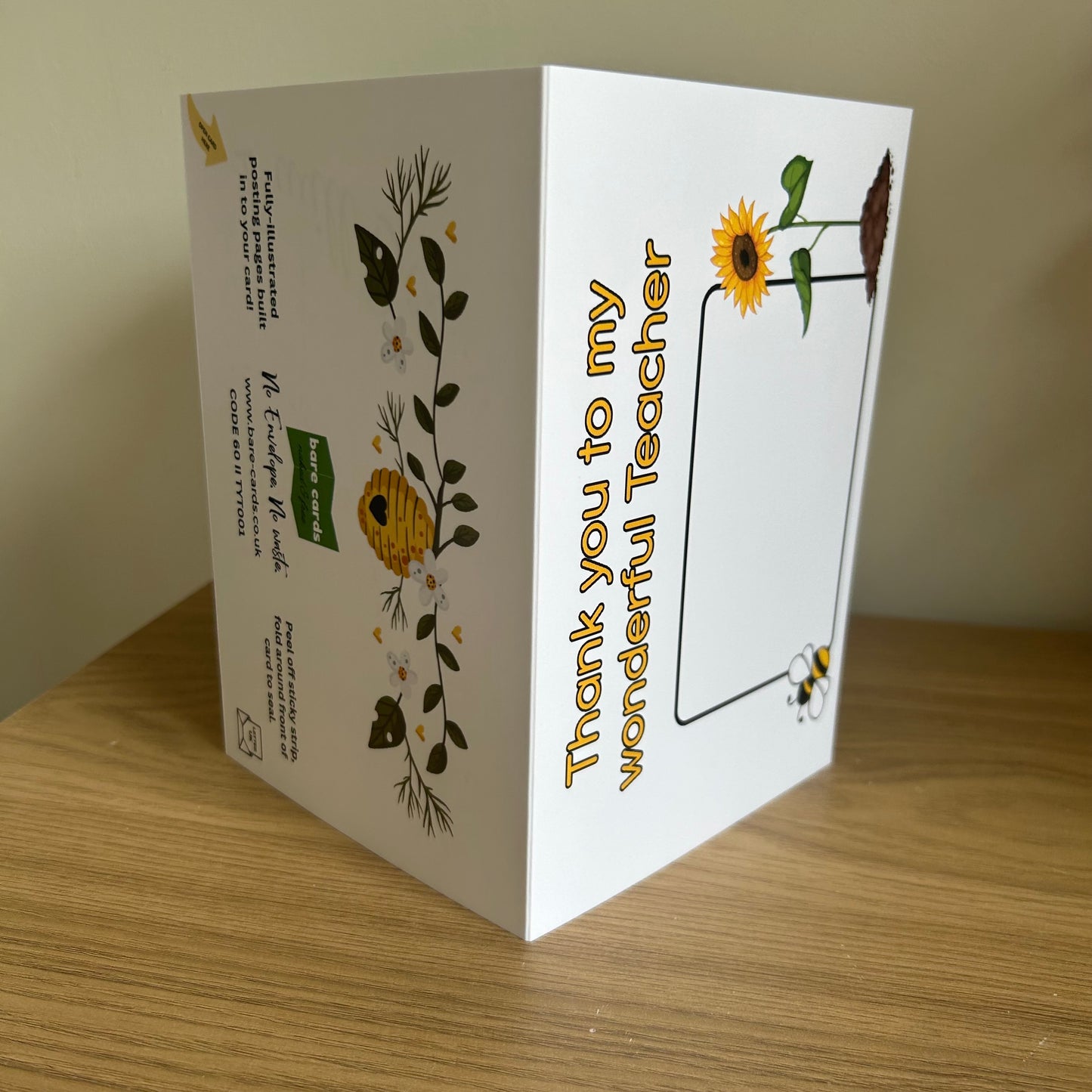 Bare Cards Thank you teacher card. Photo shows the built in envelope pages with the caption 'Thank you to my wonderful teacher' on the front with a space to write teacher name and images of a sunflower and a bee. On the back there is an image of a beehive with heart opening on a branch of white flowers. There is also the Bare Cards contact information and retail codes.
