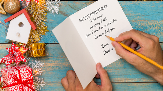 A top-down view of a person writing on a white Christmas greeting card with a yellow pencil, placed on a rustic blue wooden table. The card reads 'MERRY CHRISTMAS To the most amazing child that I could ever wish for So proud of you Love Dad x.