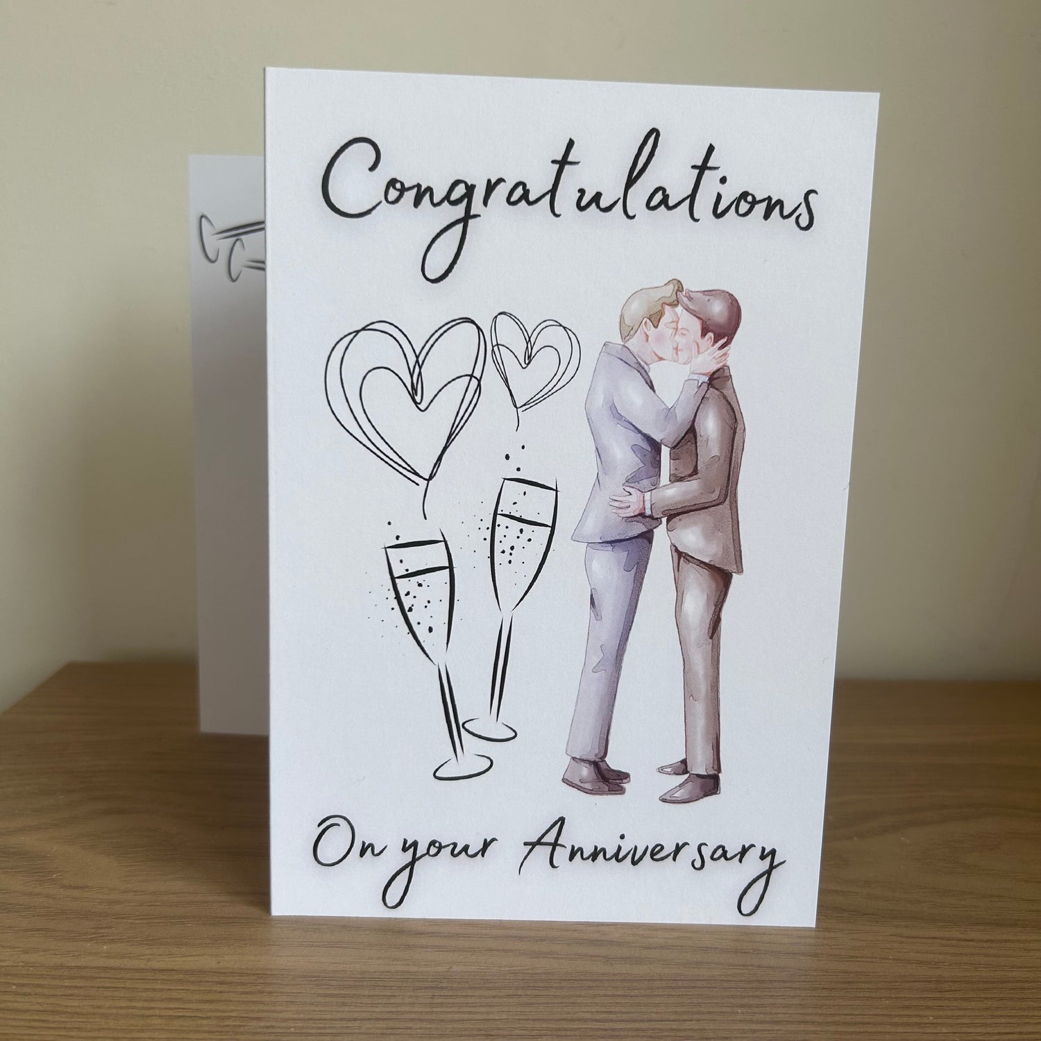 A white anniversary greeting card standing on a wooden surface, printed with the words 'Congratulations On your Anniversary' and an illustration of a male kissing couple, champagne glasses, and heart shapes. A second card is partially visible behind it.