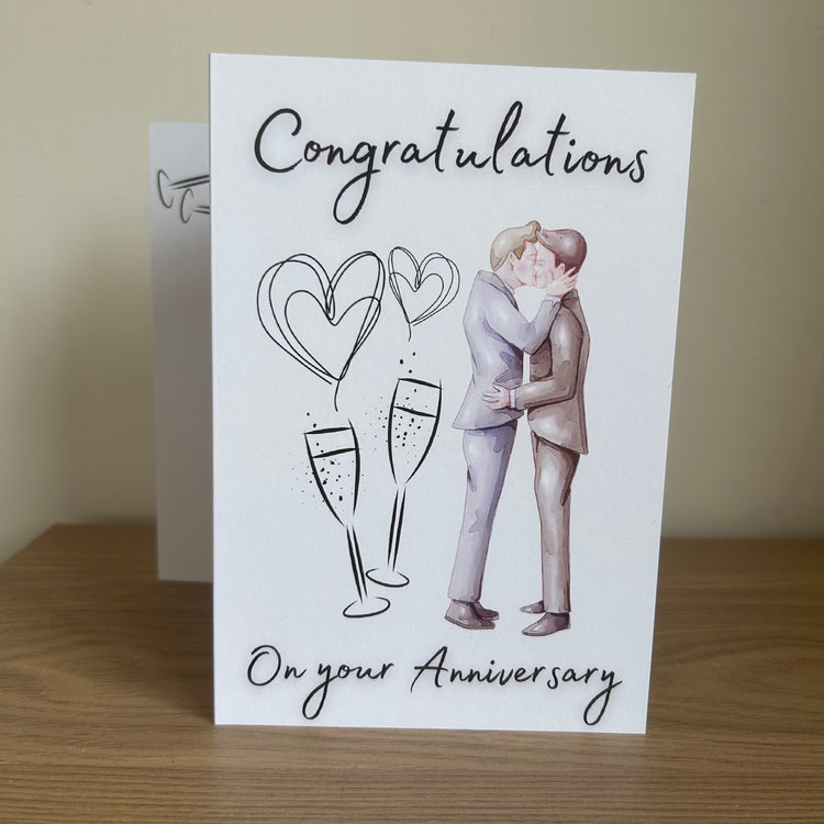 A white anniversary greeting card standing on a wooden surface, printed with the words 'Congratulations On your Anniversary' and an illustration of a male kissing couple, champagne glasses, and heart shapes. A second card is partially visible behind it.
