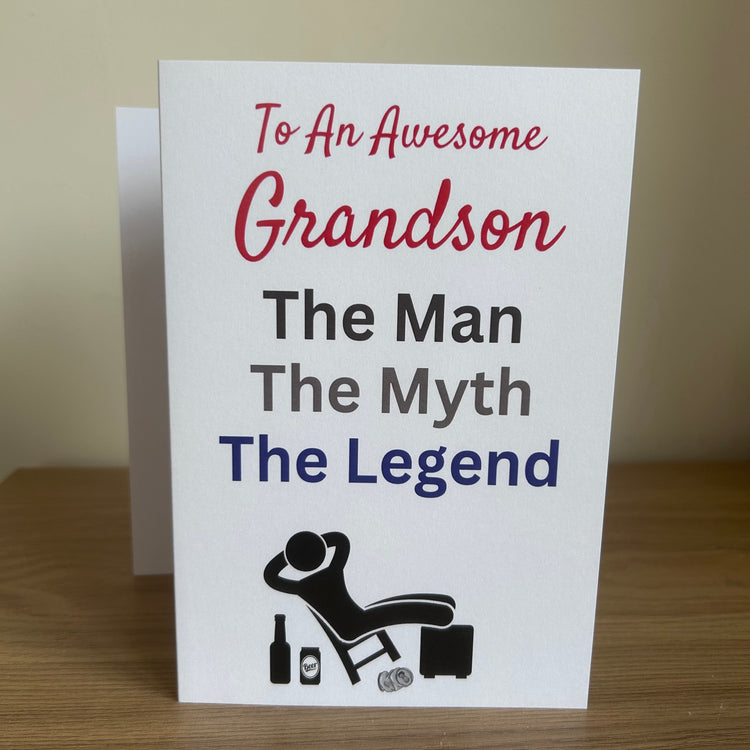 A white greeting card standing upright on a wooden surface against a beige wall, printed with the message 'To An Awesome Grandson The Man The Myth The Legend' in red, gray, and blue text, plus a small black reclining figure graphic.