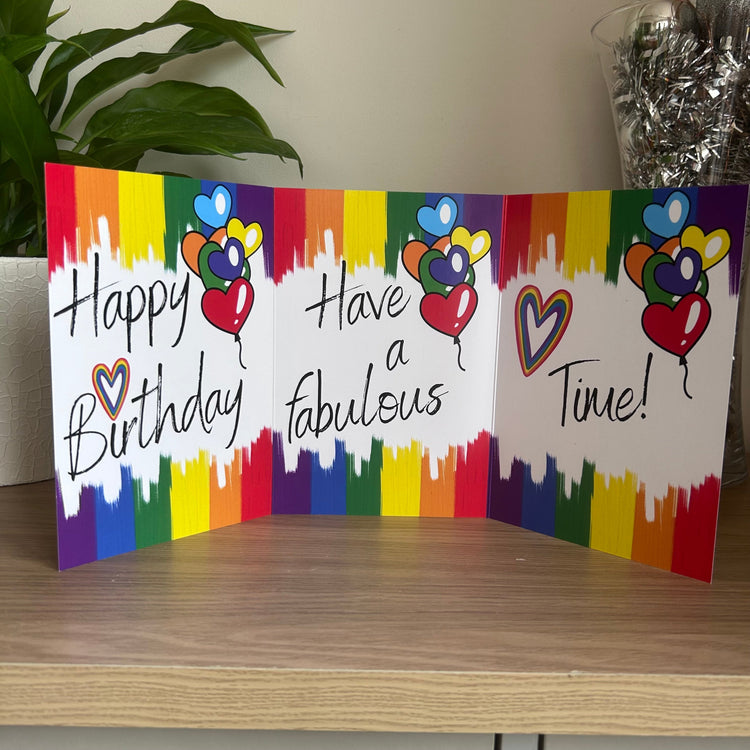 A trifold rainbow-themed birthday card standing open on a wooden table, featuring the text 'Happy Birthday', 'Have a fabulous Time!', and colorful balloon illustrations against a white background with rainbow paint splatters.