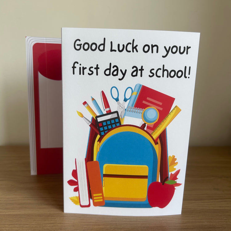 A greeting card standing on a wooden surface, with a red envelope behind it. The card reads 'Good Luck on your first day at school!' above a colorful illustration of a school backpack filled with books, pencils, scissors, and other school supplies.