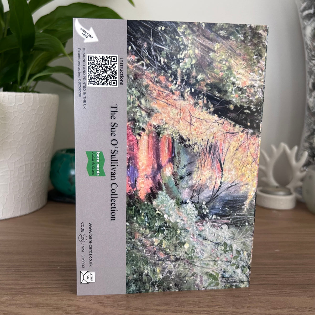 A photo of the back of the posting cover for  the self-sealing greetings card Autumn Leaves from 'The Sue O'Sullivan Collection' featuring a landscape painting of an autumn forest with yellow, green, and orange foliage on the right, and a beige sidebar on the left containing text, a QR code, and a green 'Bare Cards' logo.
