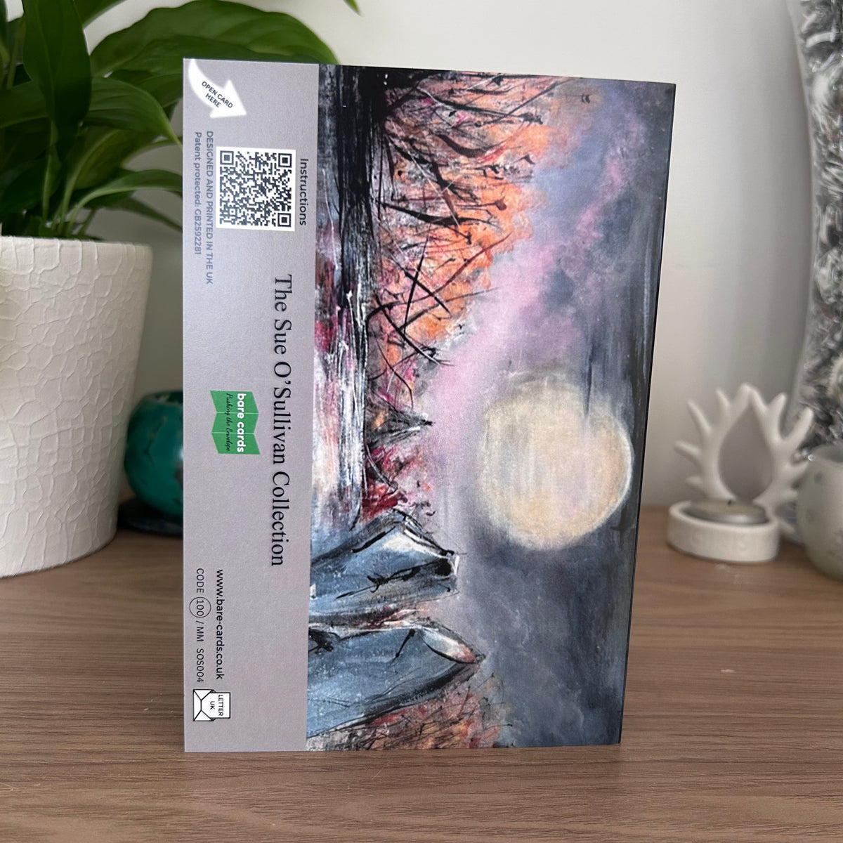 The back panel of a self-sealing greetings card featuring a moonlit landscape painting with a large pale moon, dark silhouetted trees, and pink-gray sky, displayed standing on a wooden surface. The left side has a gray printed border with text reading 'The Sue O'Sullivan Collection' and a green Bare Cards logo.