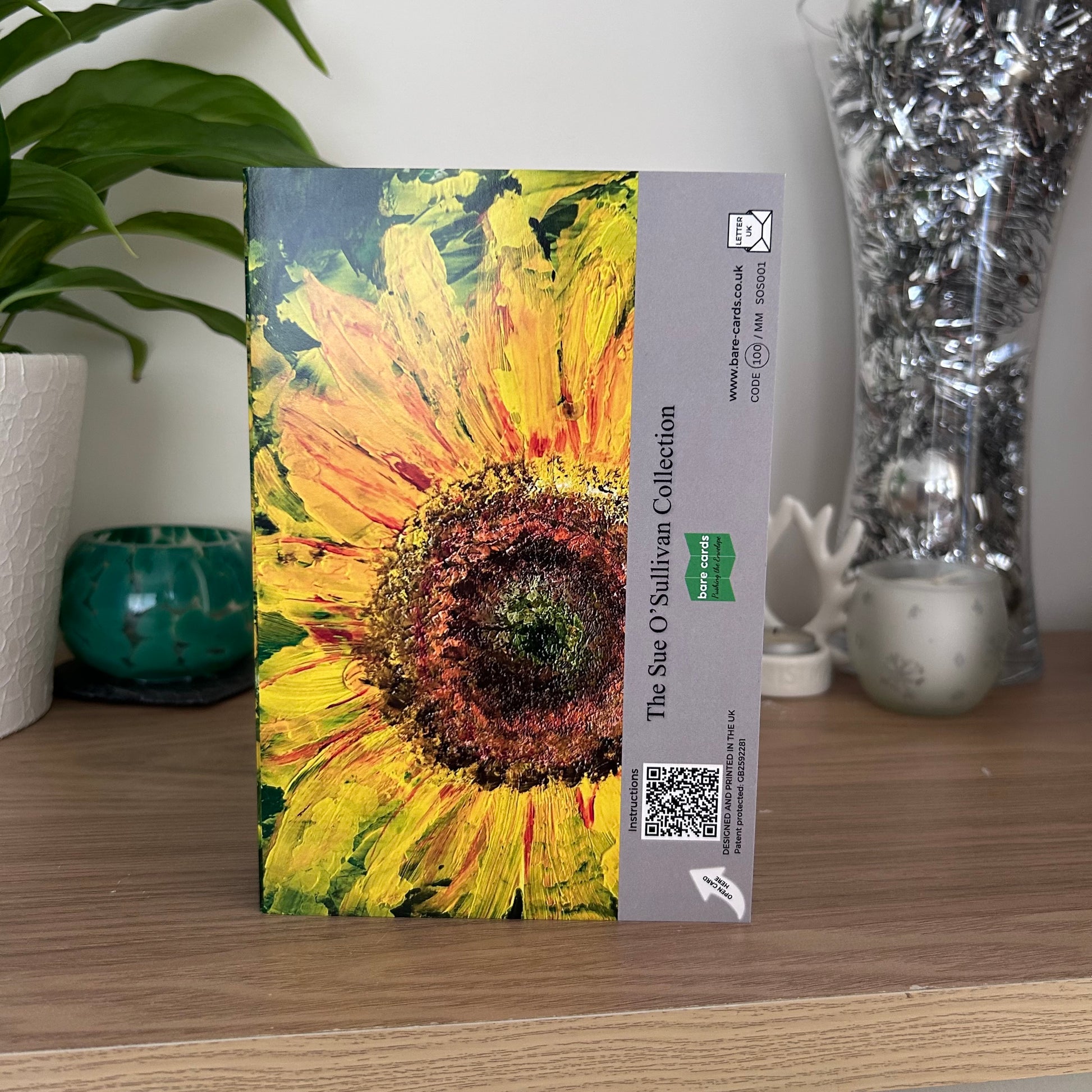 A photo featuring the back envelope panel of the self-sealing greetings card showing an abstract textured painting of a sunflower, with a beige side panel containing text, a QR code, and a green Bare Cards logo. The text reads 'The Sue O'Sullivan Collection'.
