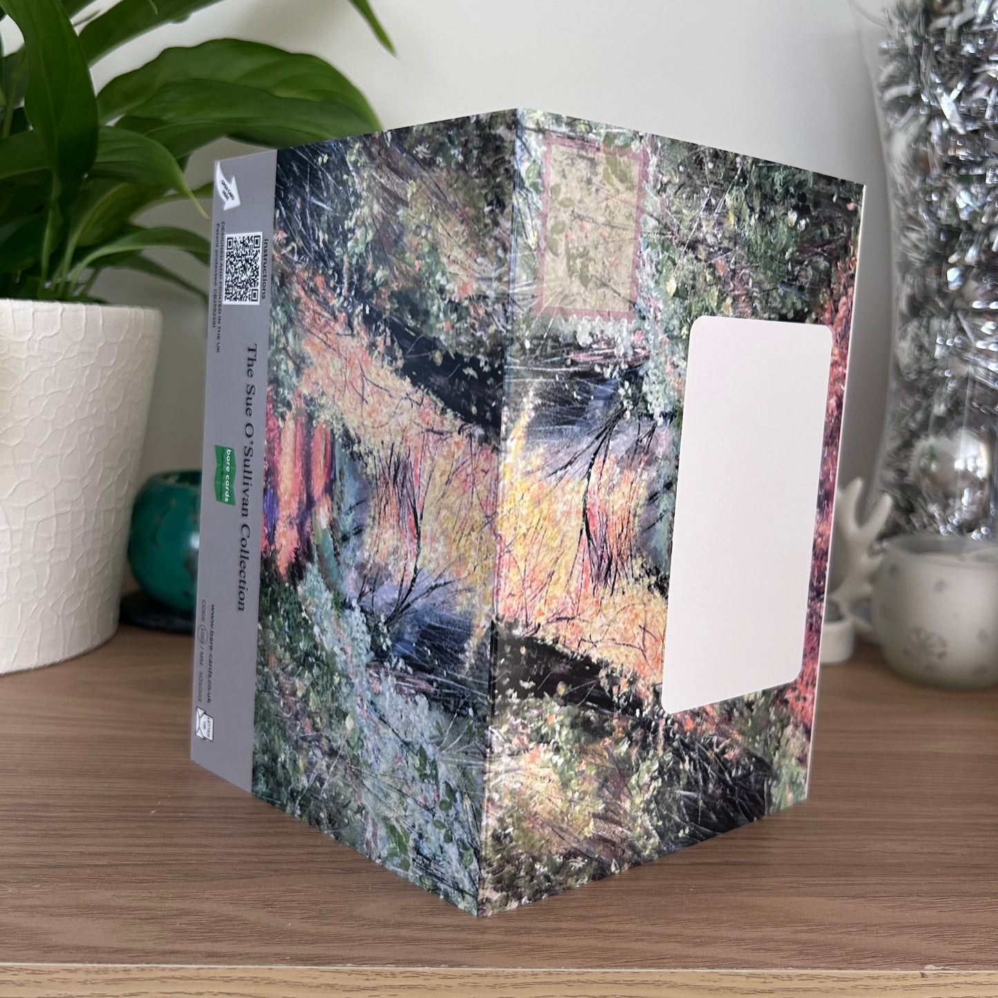 The posting covers of the Bare Cards Self-sealing greetings card - Autumn Trees, displayed on a wooden tabletop with a potted plant and decorative silver vase in the background. The print features a n autumn forest painting on both sides and product information and brand logo on the back