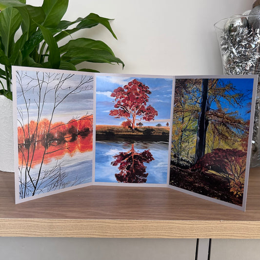 A three-panel folding art greetings card displaying autumn landscape scenes with tree reflections in water, photographed open on a wooden surface with a plant and decorative container in the background.