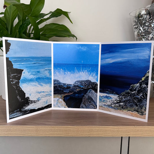 A three-panel folding art greetings card displaying coastal ocean scenes, displayed on a wooden tabletop with a green plant in the background.