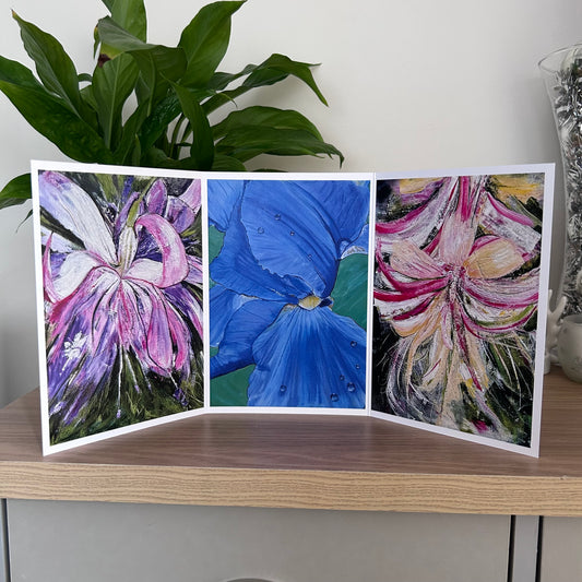 A three-panel folding art greetings card featuring colorful abstract floral paintings, displayed on a wooden shelf with a green plant in the background.
