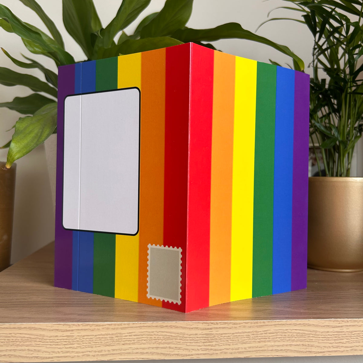 A greetings card standing on a wooden shelf showing rainbow striped posting covers, with a white address label on the front and a small beige stamp-shaped label on the side. Green potted plants are visible in the background.