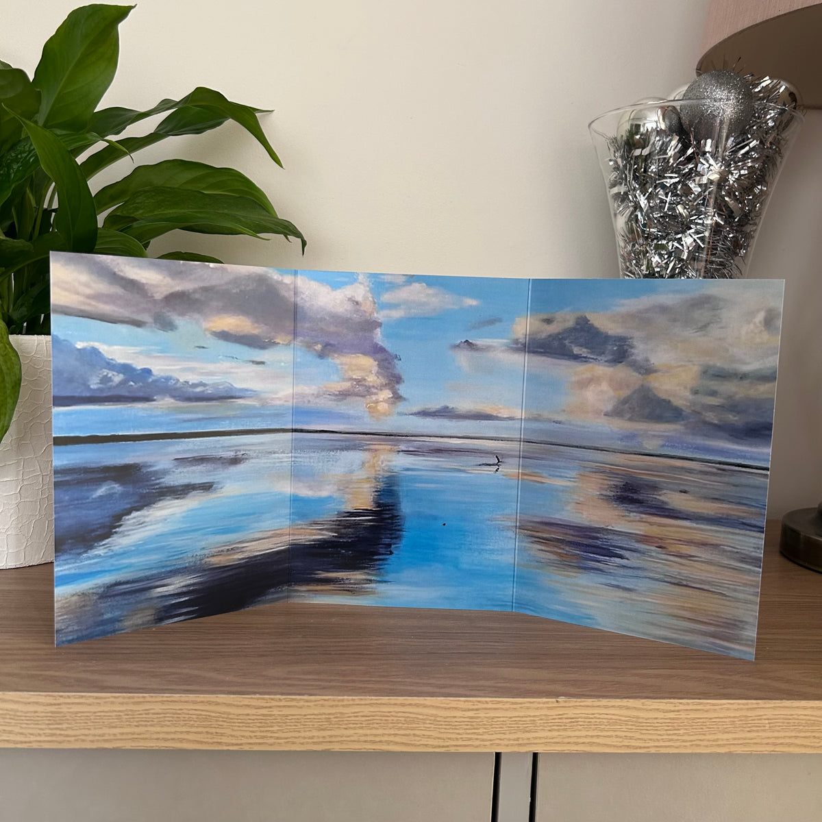 A three-panel folding greetings card featuring a painted seascape with blue sky, white and gray clouds, and reflections in the water, displayed on a wooden shelf next to a green plant.