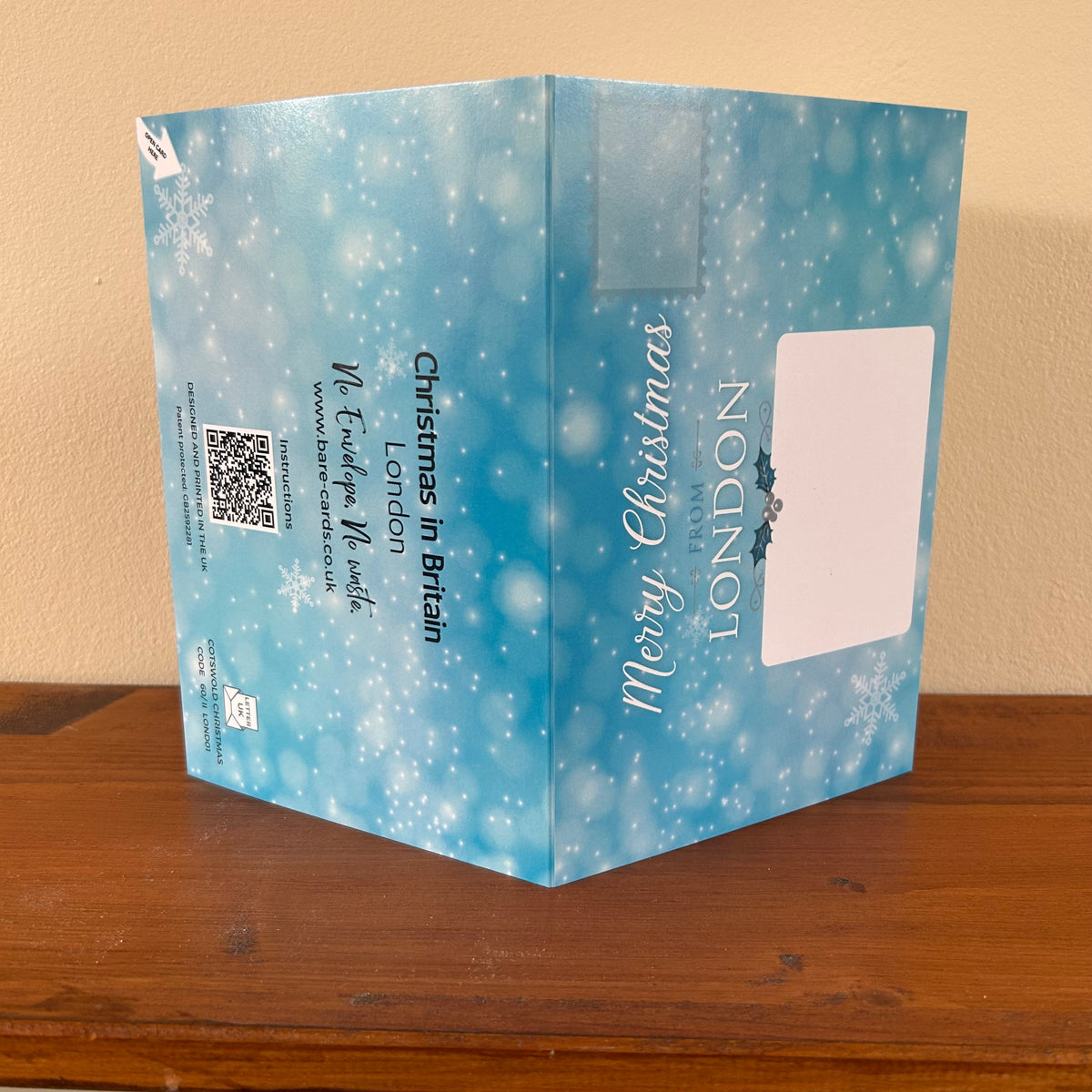 Bare Cards self-sealing posting pages for the London Christmas Cards with white snowflake on a pale blue background, featuring the text 'Merry Christmas from LONDON' and 'Christmas in Britain London', displayed on a wooden surface against a beige wall.