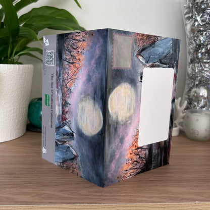 The posting covers of the Bare Cards Self-sealing greetings card - Moonlit Henge, displayed on a wooden tabletop with a potted plant and decorative silver vase in the background. The print features a moody, painterly night scene with two glowing full moons, dark silhouetted trees, and a purple and orange sky.