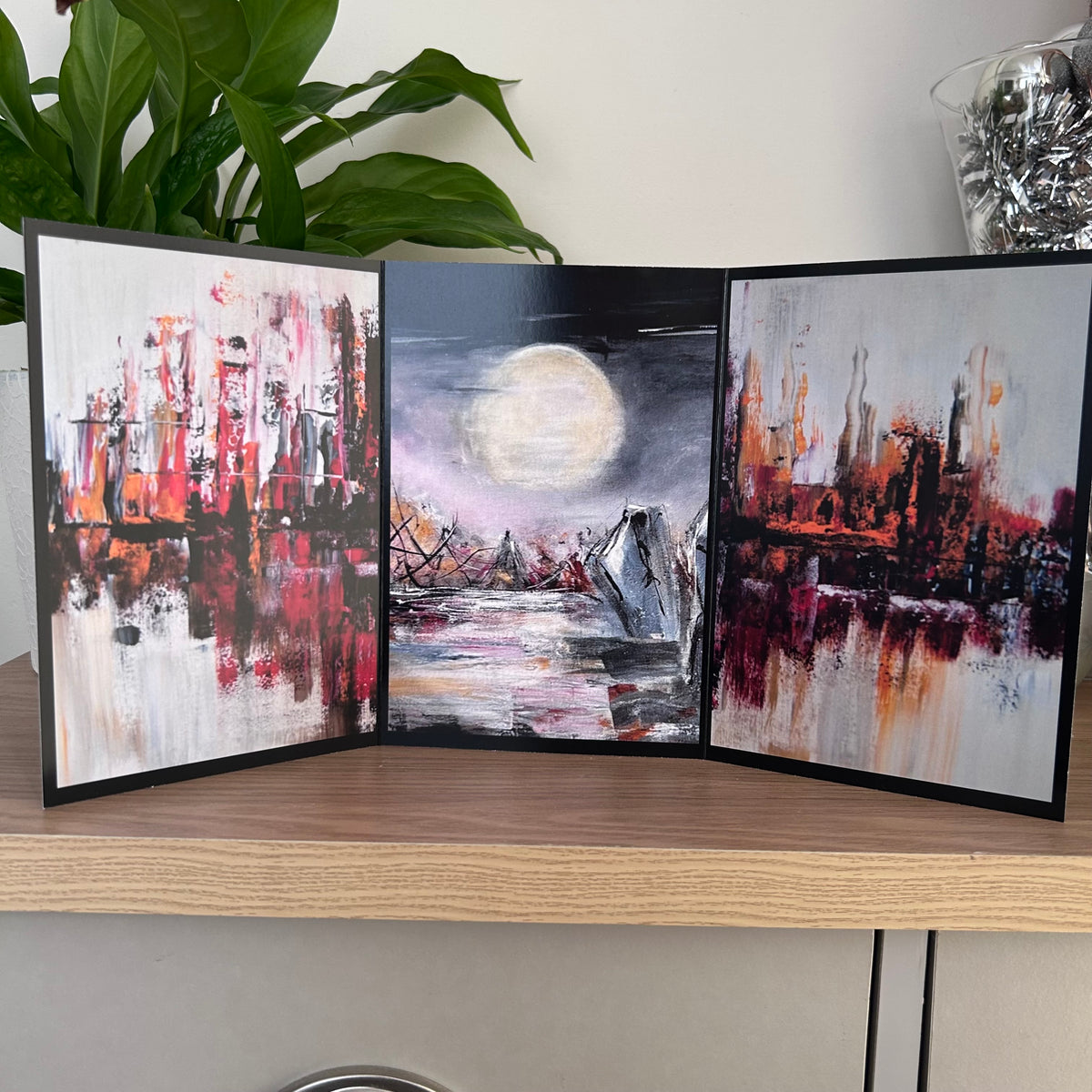 A three-panel art greetings card featuring an abstract cityscape with red, black, and white brushstrokes, a central night scene with a large moon over water, and another cityscape on the right panel, displayed on a wooden shelf.