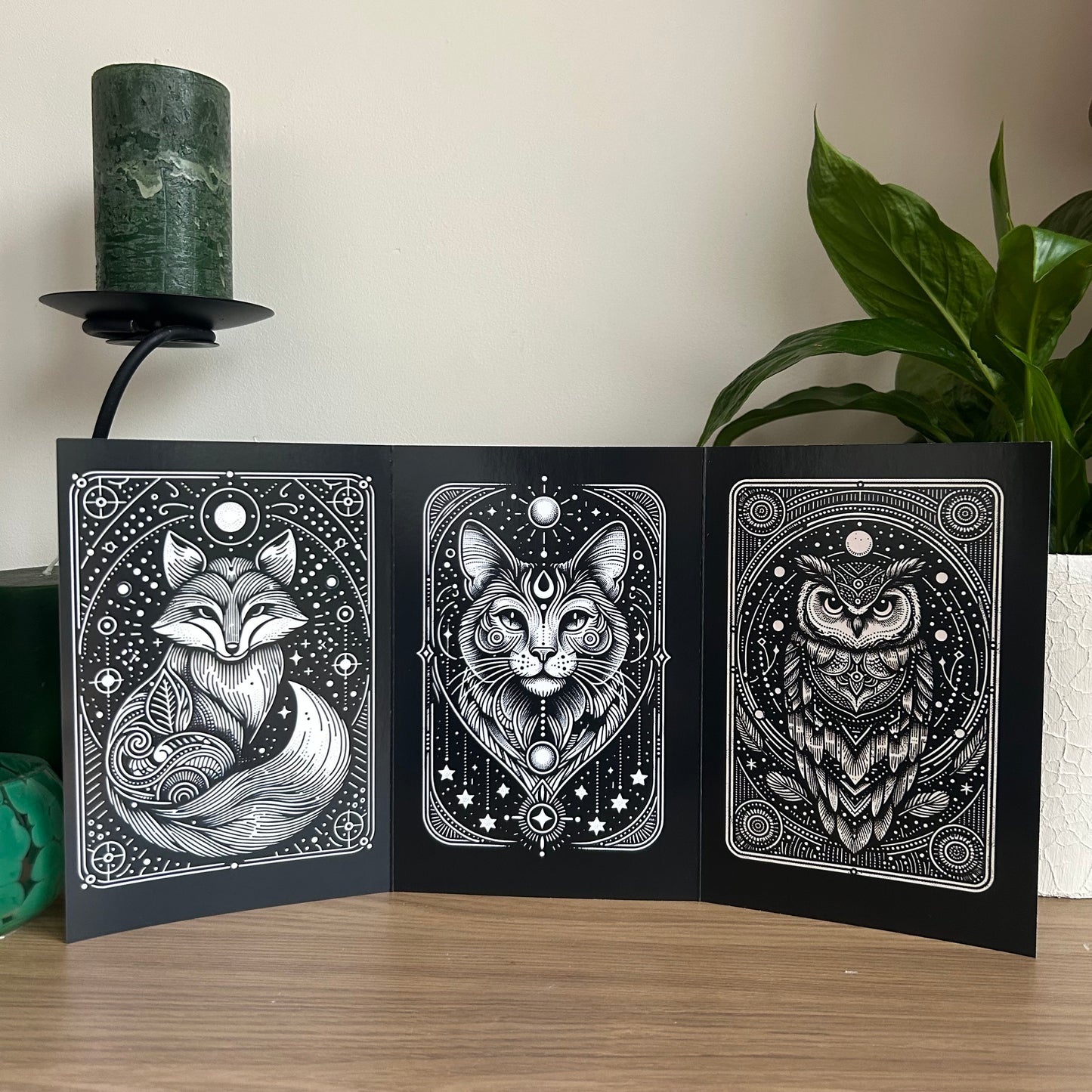 Three black and white illustrations of a fox, cat, and owl on a wooden surface with a plant and candle in the background.