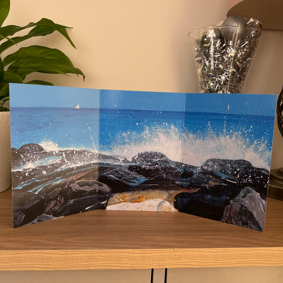 A three-panel trifold greetings card of a rocky coastline with crashing blue waves, displayed on a wooden table in a home setting.