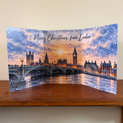 A Christmas greeting card featuring a festive London winter scene with Big Ben, Westminster Bridge, and snow falling under a warm sunset sky. The card is displayed standing on a wooden surface against a beige wall, with the text 'Merry Christmas from London' printed across the top.