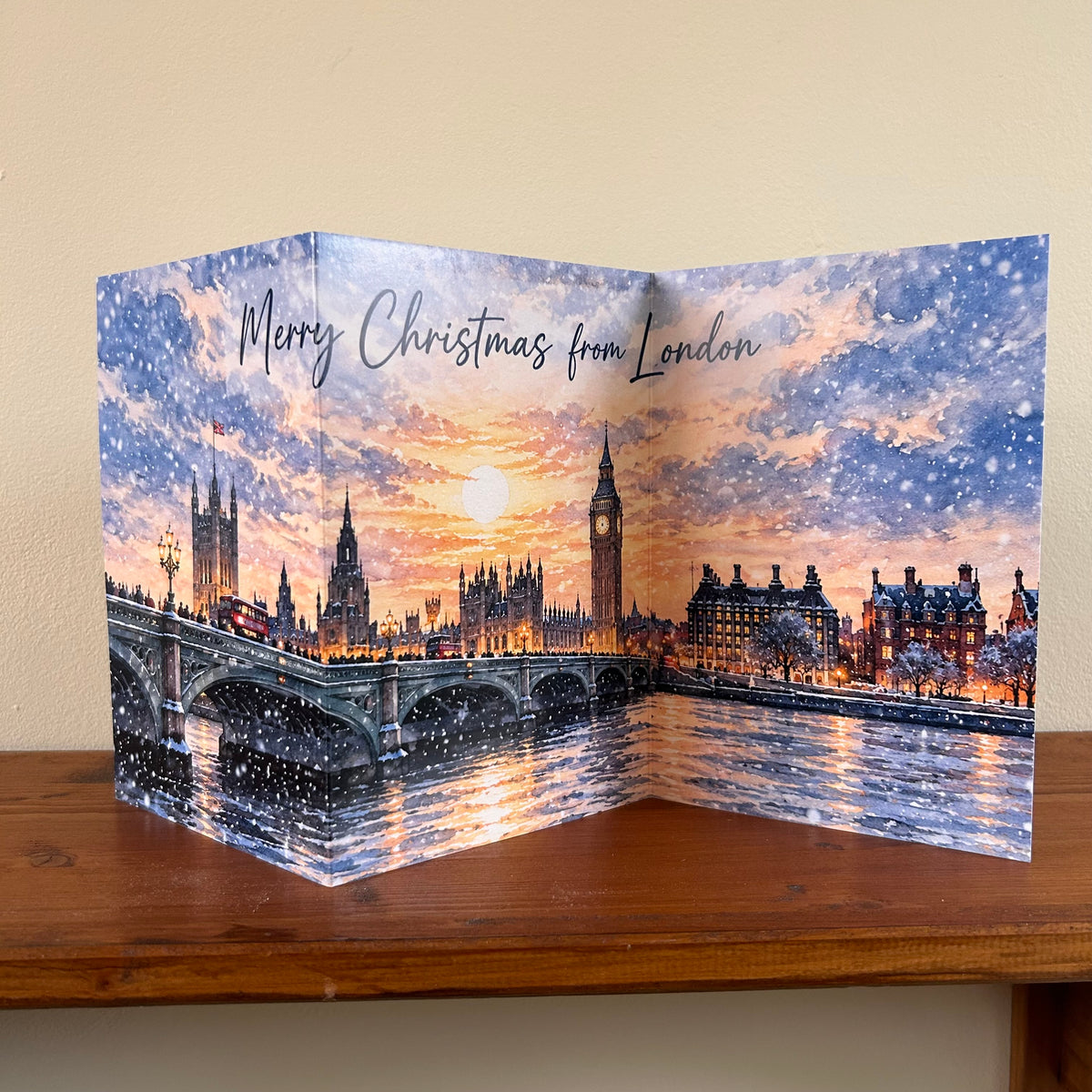 A self-sealing Christmas greeting card featuring a festive London skyline with Big Ben, Westminster Bridge, and red buses under a snowy, glowing sky. The card is displayed standing on a wooden shelf against a beige wall, with 'Merry Christmas from London' written at the top.