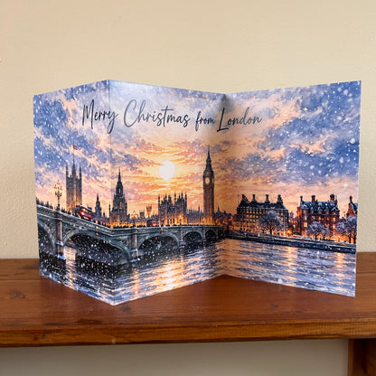 A self-sealing Christmas greeting card featuring a festive London skyline with Big Ben, Westminster Bridge, and red buses under a snowy, glowing sky. The card is displayed standing on a wooden shelf against a beige wall, with 'Merry Christmas from London' written at the top.