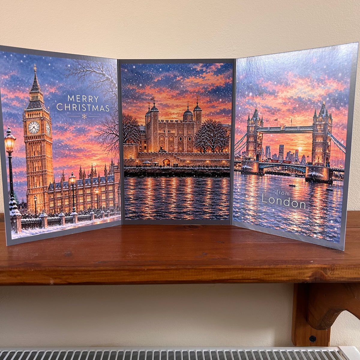A three-panel folding Christmas card featuring London landmarks, displayed on a wooden shelf against a beige wall. The left panel shows Big Ben at Christmas with 'Merry Christmas' text, the center panel shows Tower Hill with a sunset sky, and the right panel shows Tower Bridge with 'To You London' text.