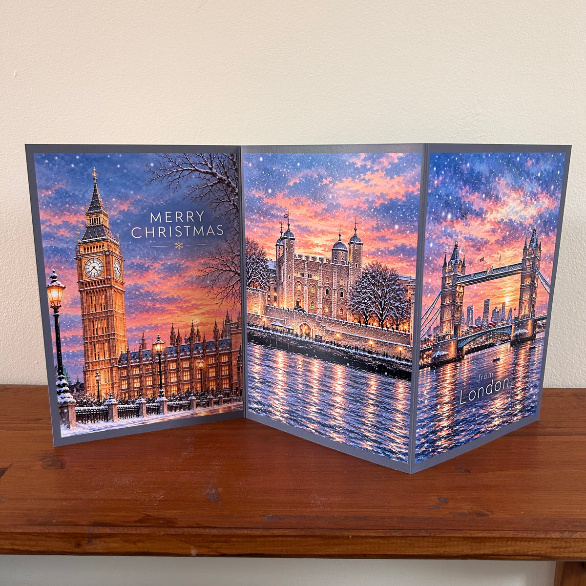 A three-panel Christmas greeting card featuring London landmarks including Big Ben, the Tower of London, and Tower Bridge in a snowy winter sky, displayed on a wooden surface against a beige wall.