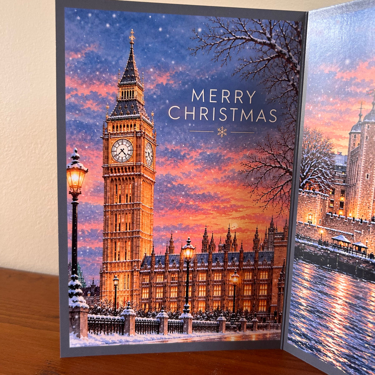 Panel one of the triptych London Christmas Card from Bare Cards on a wooden surface, showing a snowy London scene with Big Ben and the Houses of Parliament under a colorful sunset sky. The card reads “MERRY CHRISTMAS.