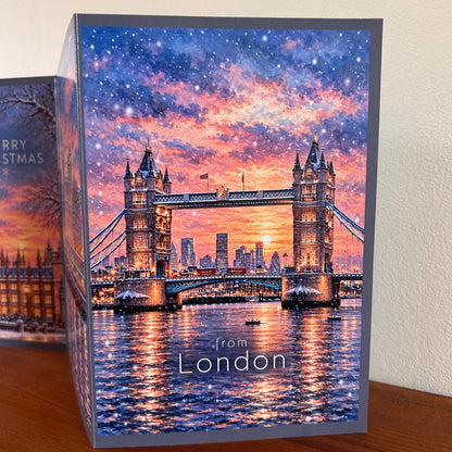 The third panel of the Bare Cards self-sealing triptych London Christmas Card featuring a London Tower Bridge sunset scene with falling snow, printed on a gray cardstock border. The card is standing on a wooden surface against a light beige wall, with the other panels partially visible behind it.