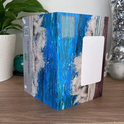 The posting panels of the self-sealing greetings cards in the Sue O'Sullivan collection with an abstract blue and white wave design, standing on a wooden table next to a potted plant.
