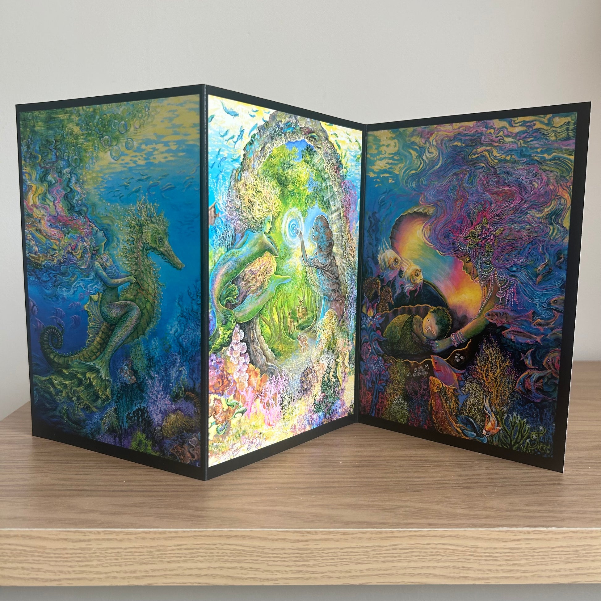 Three-panel self-sealing greetings card UK with artistic designs of under the sea and mermaid figures on a wooden surface.