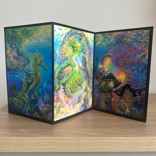 Three-panel self-sealing greetings card UK with artistic designs of under the sea and mermaid figures on a wooden surface.