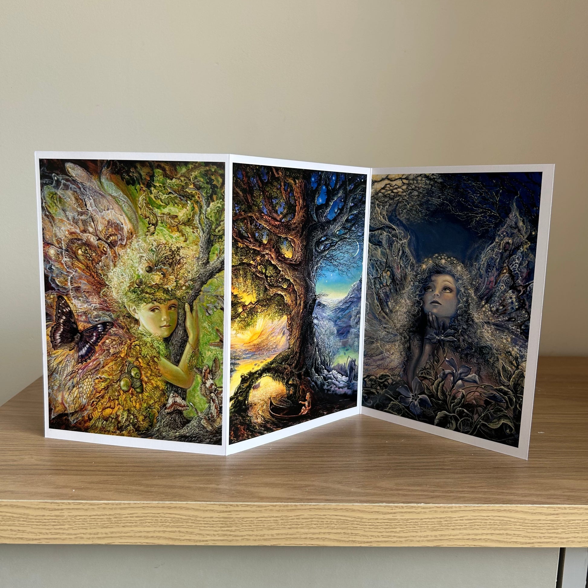 Three-panel self-sealing greetings card UK with artistic fantasy designs featuring Josephine Wall paintings of nature, fairies and mystical figures on a wooden surface