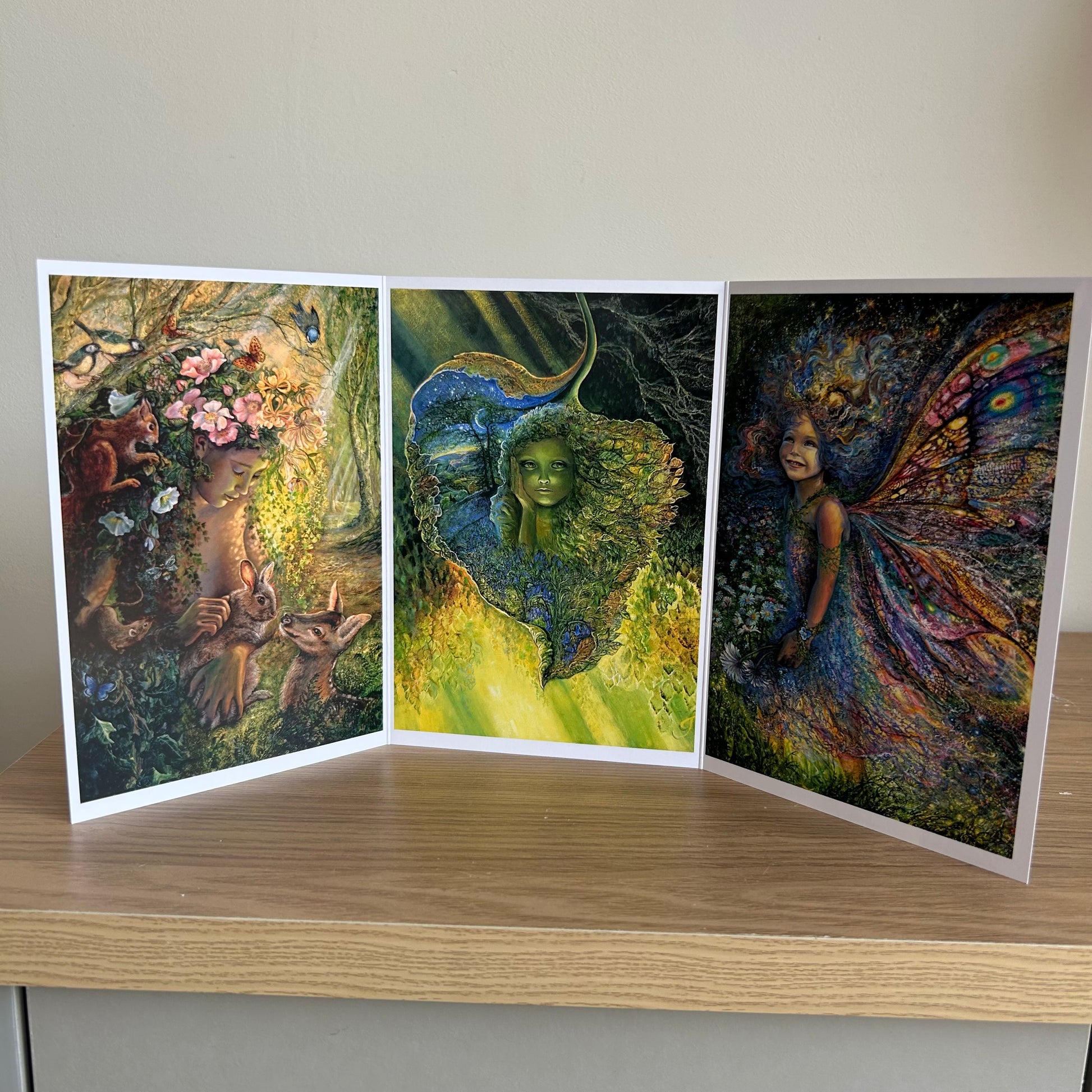 Three-panel self-sealing greetings card UK with artistic fantasy designs featuring Josephine Wall paintings of nature, fairies and mystical figures on a wooden surface