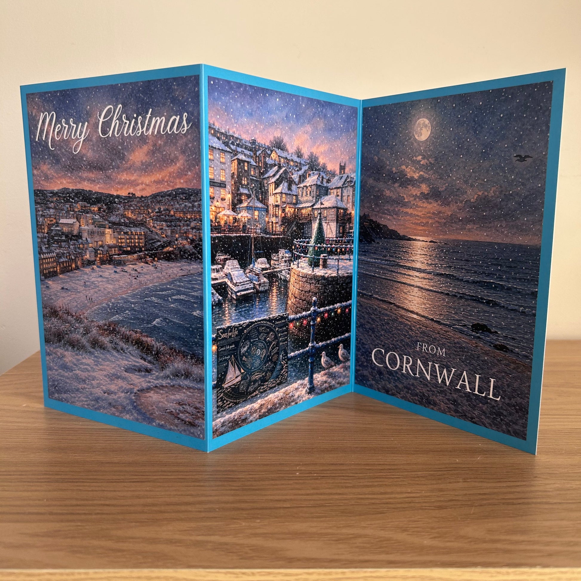 A Trifold Christmas greeting card displayed open on a wooden surface, showing a snowy coastal harbor scene on the left panel, a starry night sky over the sea on the right panel, and the text 'Merry Christmas' and 'FROM CORNWALL'.