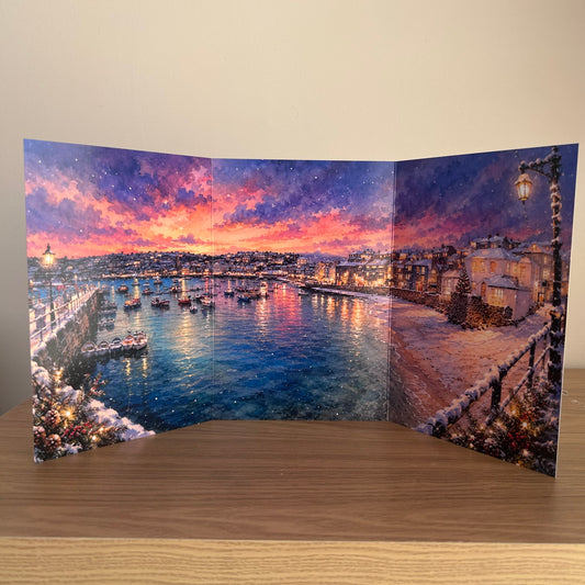 A three-panel folding Christmas Card featuring a snowy harbor sunset scene at St Ives with colorful sky, boats, and illuminated buildings, set against a plain beige wall and wooden tabletop.