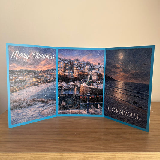 A triptych Christmas greeting card displayed open on a wooden surface against a beige wall. The card features winter coastal scenes from Cornwall, with snow-covered hills, a harbor at dusk, and a starry night sky over the sea.