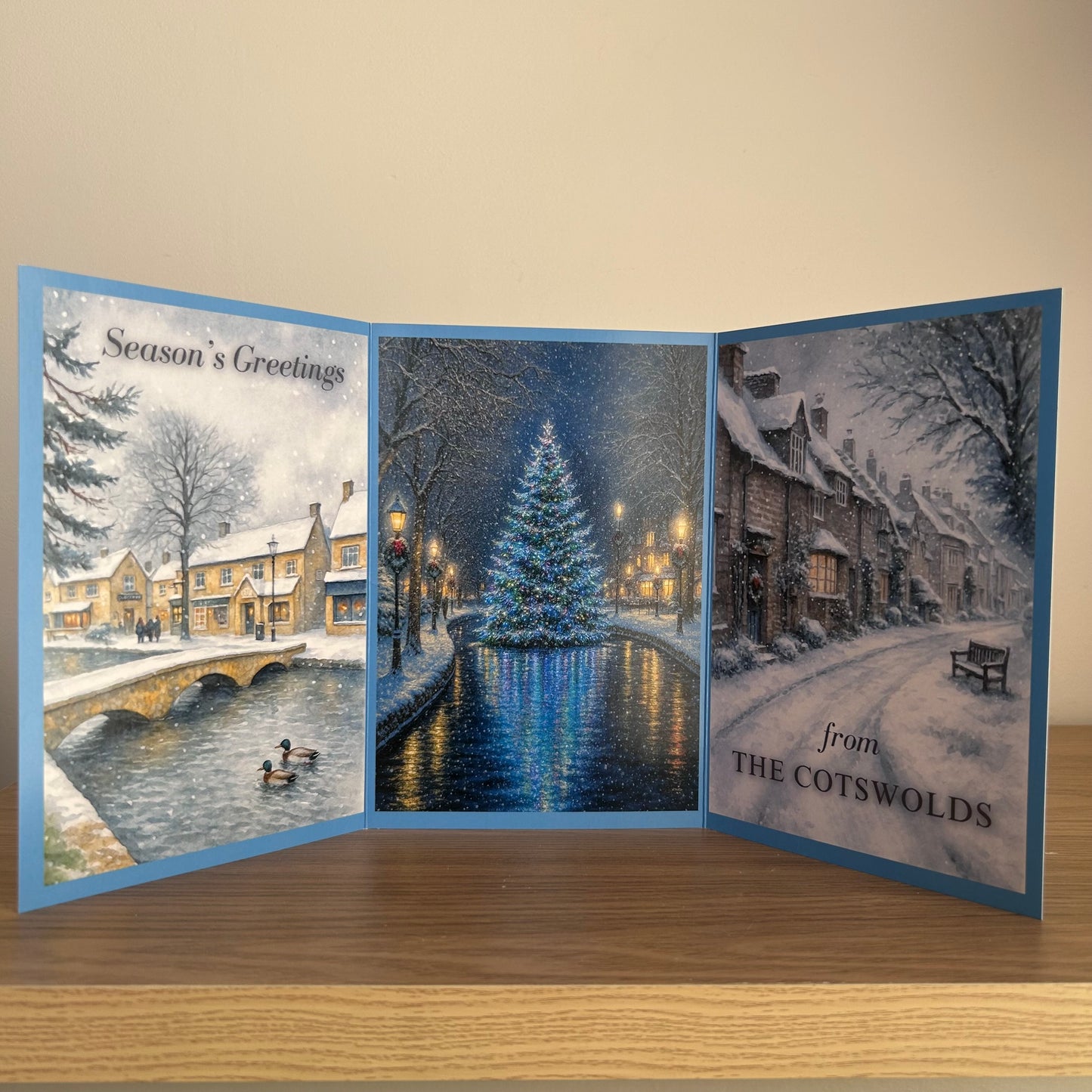 A three-panel folding Christmas greeting card displayed open on a wooden surface against a beige wall. The card features winter village scenes: a snowy bridge and cottages on the left, a glowing Christmas tree by a canal in the center, and a snow-covered street with the text 'from THE COTSWOLDS' on the right.