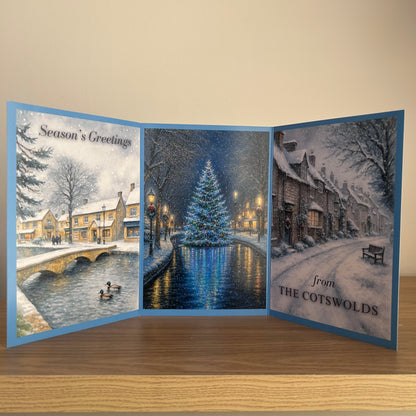 A three-panel folding Christmas greeting card displayed open on a wooden surface against a beige wall. The card features winter village scenes: a snowy bridge and cottages on the left, a glowing Christmas tree by a canal in the center, and a snow-covered street with the text 'from THE COTSWOLDS' on the right.