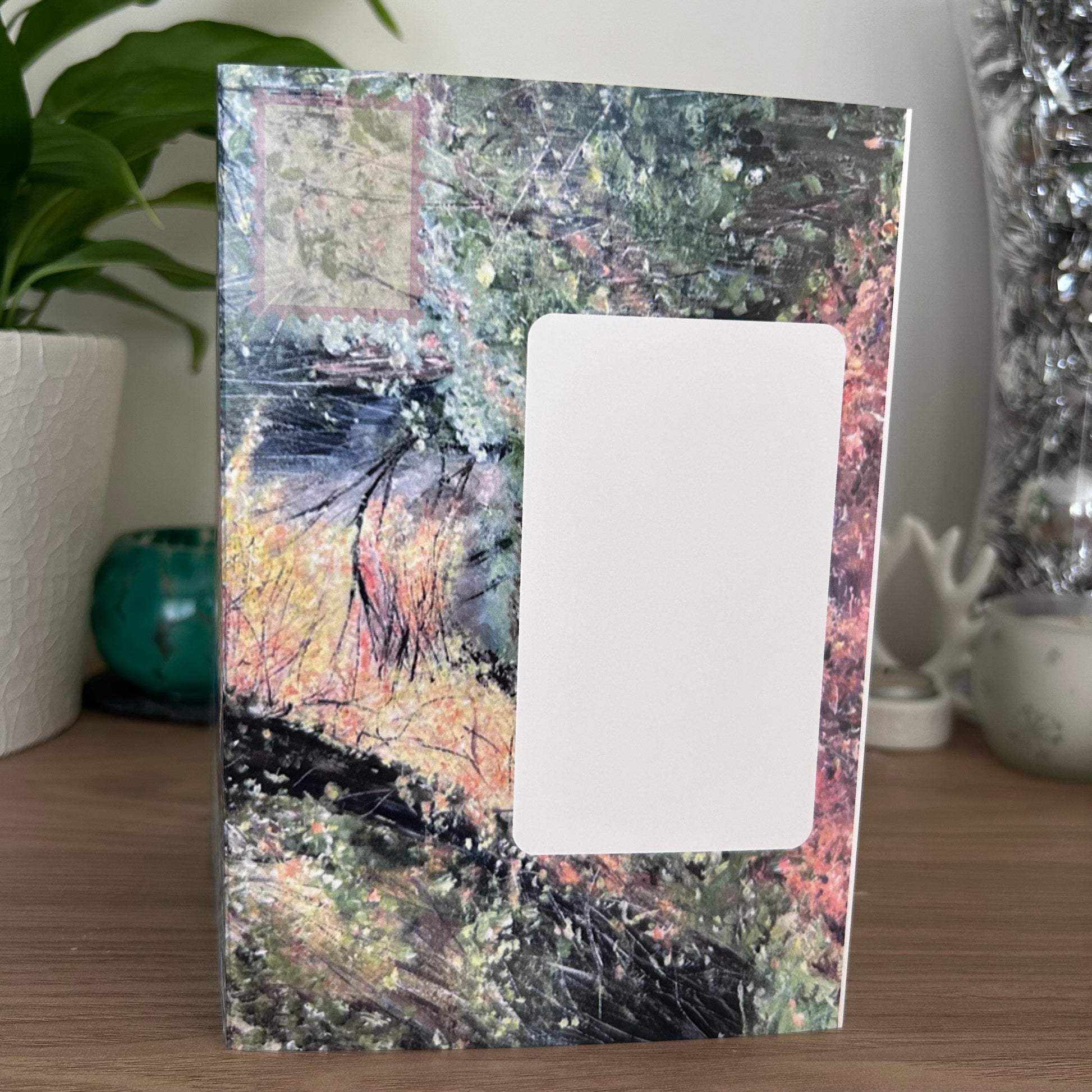 A photo of the front envelope panel of a self-sealing greetings card featuring an abstract landscape painting with a large blank white rectangular cutout in the center, displayed on a wooden surface with a potted plant and decorative items in the background.