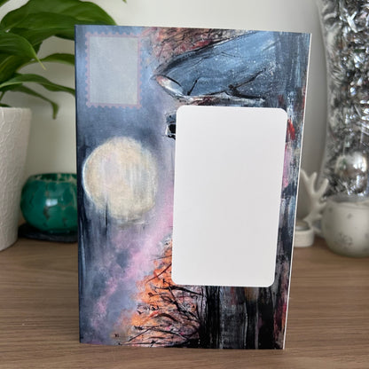 A photo of the front of the envelope panel for a self-sealing art card featuring an abstract night scene with a large white rectangular overlay in the center, displayed on a wooden surface with houseplants and decorative items in the background.