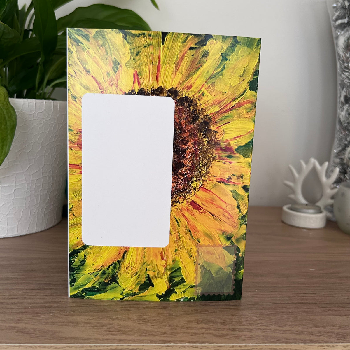 The front posting panel of a Bare Cards self-sealing greeting card featuring an impasto-style painted sunflower design in yellow, green, and brown, with a blank white rectangular photo slot on the front, standing on a wooden surface.