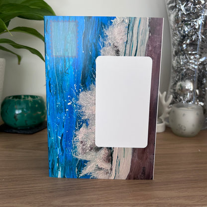 The front envelope panel of a self-sealing greetings card featuring an abstract blue and white textured design, standing upright on a wooden surface with a white placeholder block in the center, set against a home decor background.