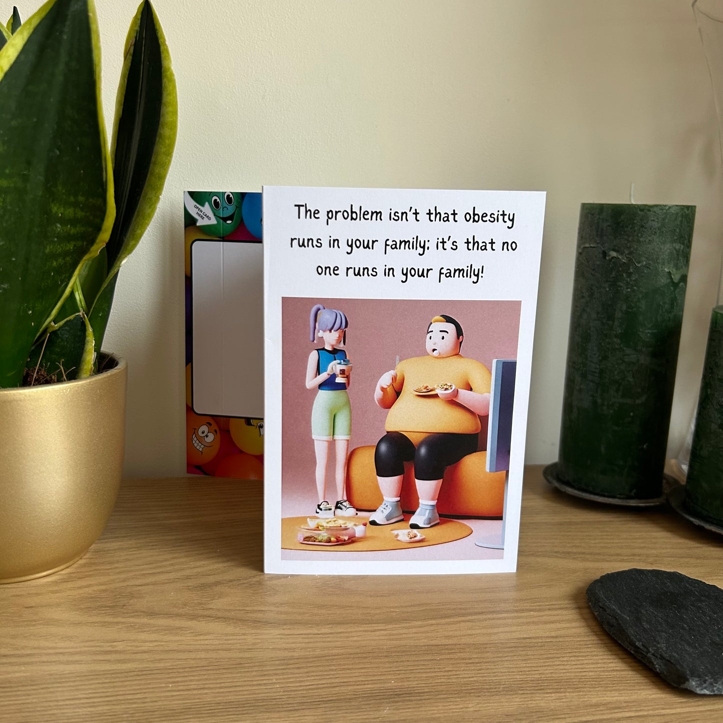 Image shows the Bare Cards Giggle Balls Humour Range 'Obesity Joke' Birthday Card displayed in a home setting. 
The card shows the image of a larger man sitting on the couch watching TV eating junk food and a slimmer female drinking coffee stood up next to him in gym wear. The caption reads "The problem isn't that obesity runs in your family: it's that no one runs in your family!"